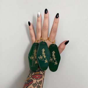 May include: Three green keychains with gold key rings and a gold design of a hand holding a plant.