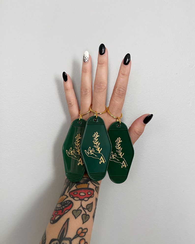 May include: Three green keychains with gold key rings and a gold design of a hand holding a plant.