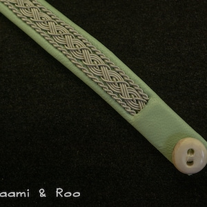 May include: A light green leather bracelet with a silver braided trim and a white button.