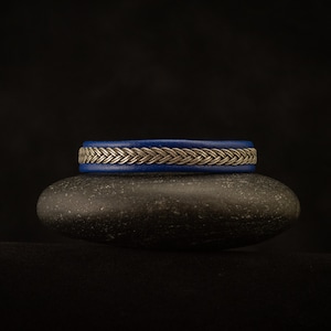 May include: A blue leather bracelet with a silver braided design, resting on a black stone.