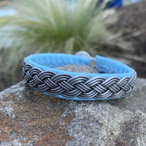Puede incluir: Un brazalete de cuero azul claro con un diseño trenzado plateado.
