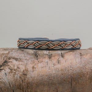 May include: A braided bracelet with a dark blue band and a woven pattern of copper and silver-colored threads. The bracelet is displayed on a piece of weathered wood, showcasing its handcrafted design.