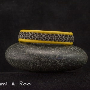 May include: A yellow leather bracelet with a braided silver and black design. The bracelet is resting on a gray stone.