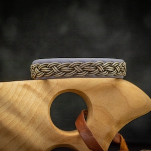 Può includere: Un braccialetto in pelle intrecciata argento e viola con un design intrecciato.