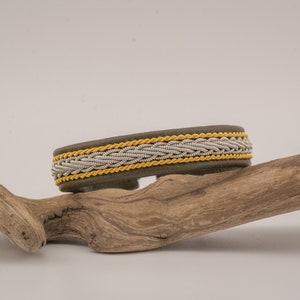 May include: A grey leather bracelet featuring a central braided silver wire pattern flanked by two parallel yellow cord accents. The bracelet has a smooth, matte finish and a structured, rounded band design that sits horizontally on a piece of wood.