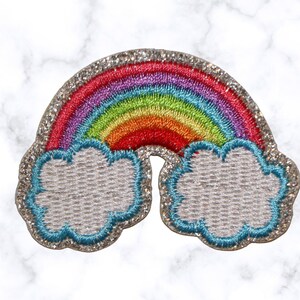Könnte beinhalten: Ein gestickter Patch mit einem Regenbogen auf silbernem Glitzer-Hintergrund. Der Regenbogen ist rot, orange, gelb, grün, blau und lila. Zwei weiße Wolken mit blauem Rand umgeben den Regenbogen.