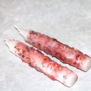 以下が含まれることがあります： 乾燥したバラの花びらとその他の植物がワックスに埋め込まれた、ピンクと白の2本のろうそく