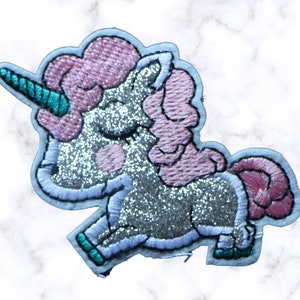 Puede incluir: Un unicornio de purpurina plateada con melena y cola rosas, bordado con puntadas blancas. El unicornio tiene un cuerno y pezuñas azul turquesa.