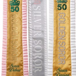 Könnte beinhalten: Drei dekorative Schärpen mit gerüschten Rändern. Links eine goldene Schärpe mit rosa Rüschen, grüner Krone, der Zahl 50, dem Text GOLDEN SOROR und Soror Debra. In der Mitte eine silberne Schärpe mit weißen Rüschen, rosa Krone, 25, SILVER SOROR und Soror Barbara. Rechts eine goldene Schärpe mit weißen Rüschen, grüner Krone, 50, GOLDEN SOROR aus Strasssteinen und Soror Darian.