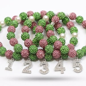 Op de afbeelding: Verschillende armbanden met groene en roze glinsterende kralen en witte parels. Elke armband heeft een zilveren bedel met een nummer van één tot vijf. De armbanden zijn gerangschikt op een witte ondergrond.