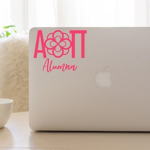 May include: A pink laptop sticker with the letters AOT and a flower design. The text "Alumma" is below the letters.