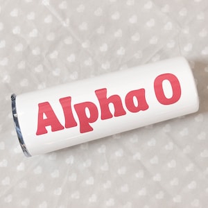 May include: White tumbler with the words "Alpha O" in red, bold, retro-style lettering. The tumbler is cylindrical and appears to be made of stainless steel. The background is a light gray fabric with a repeating heart pattern.