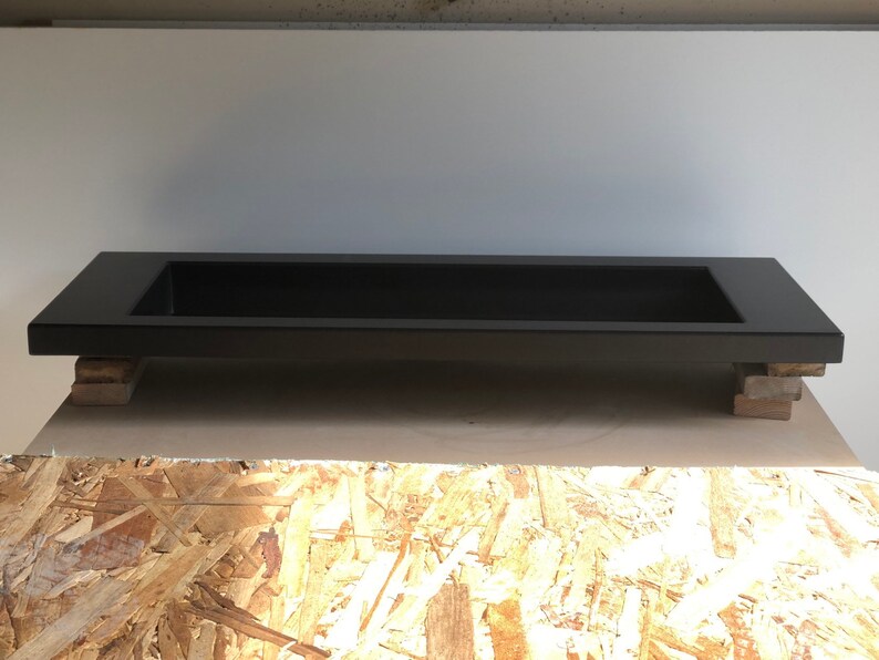 Puede incluir: Un lavabo rectangular negro con un borde elevado, apoyado en bloques de madera. El lavabo est&aacute; hecho de un material liso y s&oacute;lido.