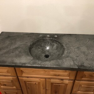 Puede incluir: Una encimera de ba&ntilde;o de concreto gris con un lavabo redondo gris y tres agujeros para grifos. La encimera est&aacute; montada en un gabinete de madera con cajones.