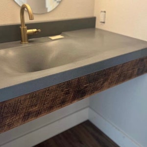 Puede incluir: Un lavabo de ba&ntilde;o de hormig&oacute;n gris con una base de madera. El lavabo est&aacute; montado en la pared y tiene un solo grifo.