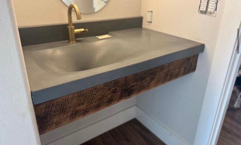 Puede incluir: Un lavabo de ba&ntilde;o de hormig&oacute;n gris con una base de madera. El lavabo est&aacute; montado en la pared y tiene un solo grifo.