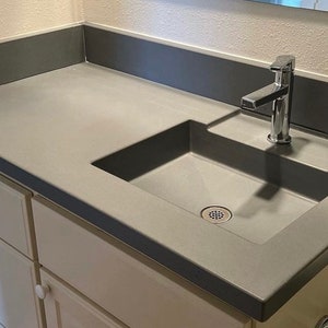Puede incluir: Un lavabo de ba&ntilde;o de encimera de concreto gris con un lavabo de porcelana blanca y un grifo cromado.