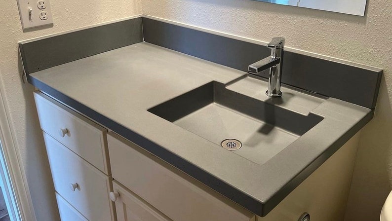 Puede incluir: Un lavabo de ba&ntilde;o de encimera de concreto gris con un lavabo de porcelana blanca y un grifo cromado.