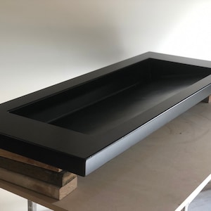 Puede incluir: Un lavabo rectangular de hormig&oacute;n negro con un borde elevado y un recorte rectangular en el centro. El lavabo est&aacute; apoyado sobre bloques de madera.