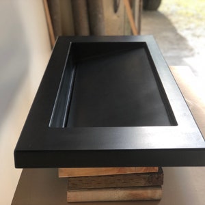Puede incluir: Un lavabo rectangular de hormig&oacute;n negro con un borde elevado y un desag&uuml;e en el centro. El lavabo est&aacute; colocado sobre una pila de tablas de madera.