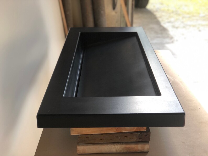 Puede incluir: Un lavabo rectangular de hormig&oacute;n negro con un borde elevado y un desag&uuml;e en el centro. El lavabo est&aacute; colocado sobre una pila de tablas de madera.