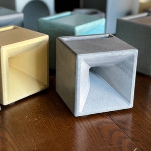 May include: A collection of modern concrete planters in various colors, including yellow, gray, and blue. The planters have a cube shape with a flared opening. The items are arranged on a wooden surface.