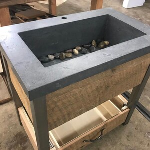 Puede incluir: Un lavabo de encimera de hormig&oacute;n gris oscuro con una base de madera y un caj&oacute;n. El lavabo tiene un peque&ntilde;o agujero de drenaje y est&aacute; lleno de piedras de r&iacute;o lisas.