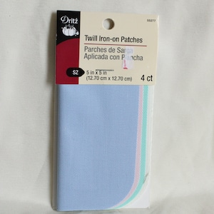 May include: A package of Dritz twill iron-on patches. The package includes four patches, each measuring 5 inches by 5 inches. The patches are light blue, with a curved edge that has pink and green stripes. The package has text in English and Spanish.