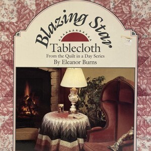 May include: A magazine cover featuring a "Blazing Star Tablecloth" design by Eleanor Burns. The image shows a round table with a patterned tablecloth, a lamp, and a red armchair. The background includes a fireplace and floral patterned fabric.