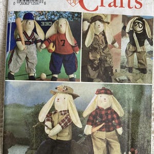 May include: Simplicity Crafts pattern featuring bunny dolls in various outfits. The bunnies are dressed in baseball uniforms, camouflage, and fishing attire. The pattern includes the text "7636" and "ONE SIZE".