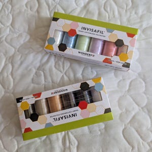May include: Two boxes of INVISAFIL Wonderfil thread spools. One box contains pastel colors, the other contains neutral colors. The boxes have a green border with colorful geometric shapes. Each spool is 400m (437 yards).