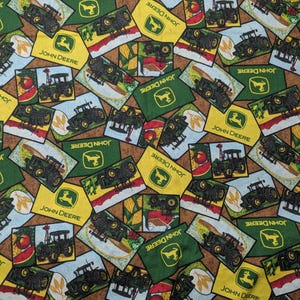May include: Fabric with a repeating pattern of John Deere tractor illustrations and logos. The design features green, yellow, and brown rectangular patches with tractors, logos, and farm scenes. The words "JOHN DEERE" are visible.