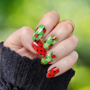 May include: Close-up of a hand with red and green nail polish. The nails are decorated with a ladybug and flower design.