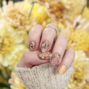 May include: Close-up of a hand with floral nail art featuring bees, honeycombs, and roses. The nails are painted in a light pink base with gold accents.