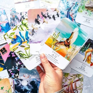May include: A set of illustrated oracle cards with various animal and nature themes. The cards feature watercolor illustrations of animals like a horse, antelope, eagle, fox, sloth, and raven. The cards are laid out on a gray surface with a hand holding one card that reads "Eagle: Ancestral Healing. Your ancestors are holding space for you to heal your past."