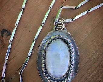 Puede incluir: Un collar de cadena de plata con un gran colgante ovalado que presenta una piedra de luna blanca engastada en un bisel de plata. El colgante tiene un acabado texturizado, martillado y un borde de cuerda trenzada.