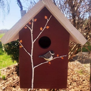 May include: A red birdhouse with a white roof and a white painted tree with orange leaves. A small bird is perched on a branch of the tree.