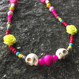 May include: A colorful beaded necklace with pink ribbon ties, white skull beads, yellow flower charms, and red, blue, yellow, and white beads.
