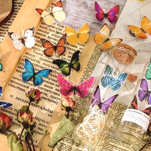May include: Two glass jars with cork tops filled with colorful butterfly stickers. The jars are sitting on a table with a newspaper and dried flowers.