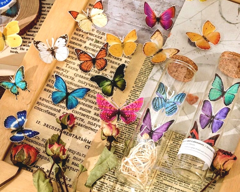 May include: Two glass jars with cork tops filled with colorful butterfly stickers. The jars are sitting on a table with a newspaper and dried flowers.