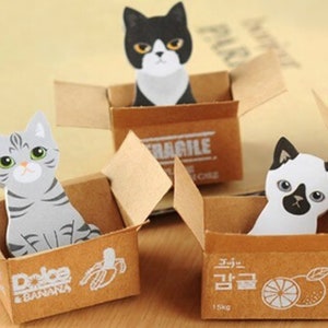 May include: Three cat-shaped paper cutouts in cardboard boxes. The cats have different color patterns: gray tabby, black and white, and white with black markings. The boxes have various printed designs, including a banana, an orange, and the word "FRAGILE".