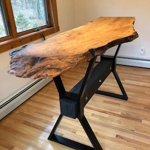 May include: A live edge wooden desk with a natural, irregular edge. The tabletop is a light brown color with darker wood grain patterns. It is supported by a black metal base with an X-shaped design. The desk is approximately 60 inches long.