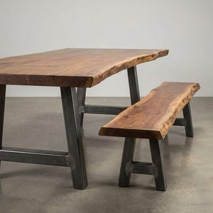 May include: A dining table and a matching bench, both featuring natural live edge wood slabs with visible grain patterns in a warm brown tone. The furniture pieces are supported by sturdy, dark gray metal A-frame legs with a smooth, industrial finish.