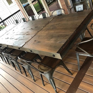 Pu&ograve; includere: Un grande tavolo da pranzo rettangolare con un piano in legno marrone scuro e una spessa struttura in metallo con bulloni visibili lungo i bordi. Il tavolo &egrave; circondato da diverse sedie in metallo con sedute in legno scuro su una terrazza.