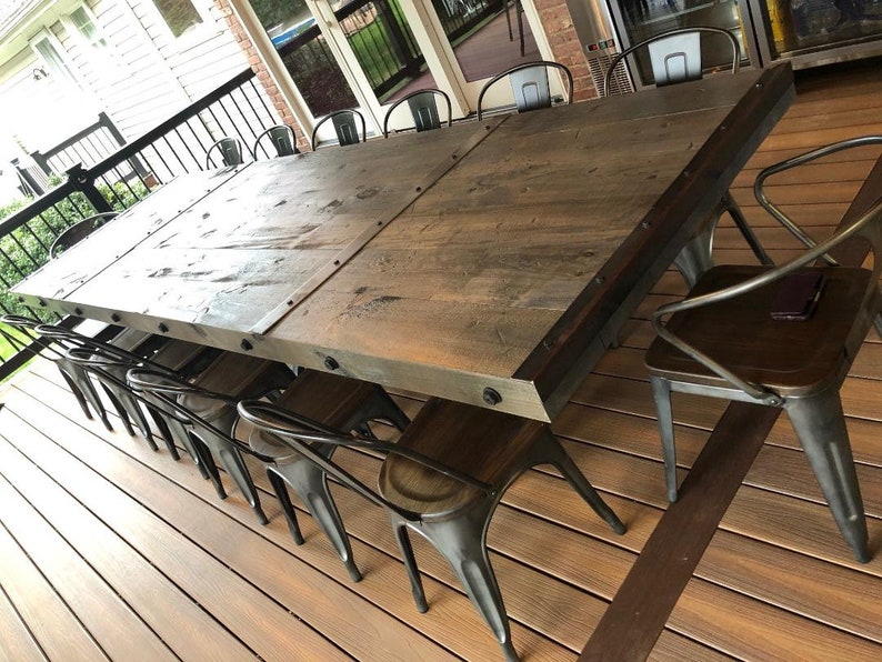 Pu&ograve; includere: Un grande tavolo da pranzo rettangolare con un piano in legno marrone scuro e una spessa struttura in metallo con bulloni visibili lungo i bordi. Il tavolo &egrave; circondato da diverse sedie in metallo con sedute in legno scuro su una terrazza.
