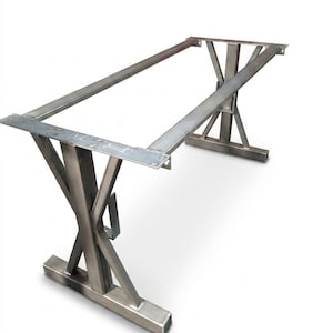 May include: A rectangular table base constructed from raw, unfinished steel. The frame features two X-shaped supports at each end, connected by two long horizontal beams. The metal surface shows visible weld marks and industrial gray tones.