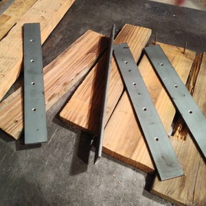 May include: Five metal strips with holes drilled along their length, lying on top of wooden planks. The metal strips are a dark gray color.