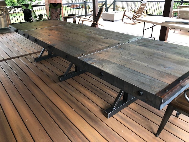 Pu&ograve; includere: Un lungo tavolo da pranzo rettangolare con un piano in legno tinto scuro composto da pi&ugrave; assi. Il tavolo &egrave; rinforzato con bordi in metallo nero fissati con bulloni a vista. Poggia su tre basi a cavalletto in metallo nero.