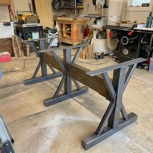 Pu&ograve; includere: Un set di due basi per tavolo realizzate in metallo grigio scuro e legno tinto marrone. Ogni base presenta una struttura di supporto centrale in metallo a forma di X collegata a una trave di legno orizzontale, con piastre metalliche piatte.