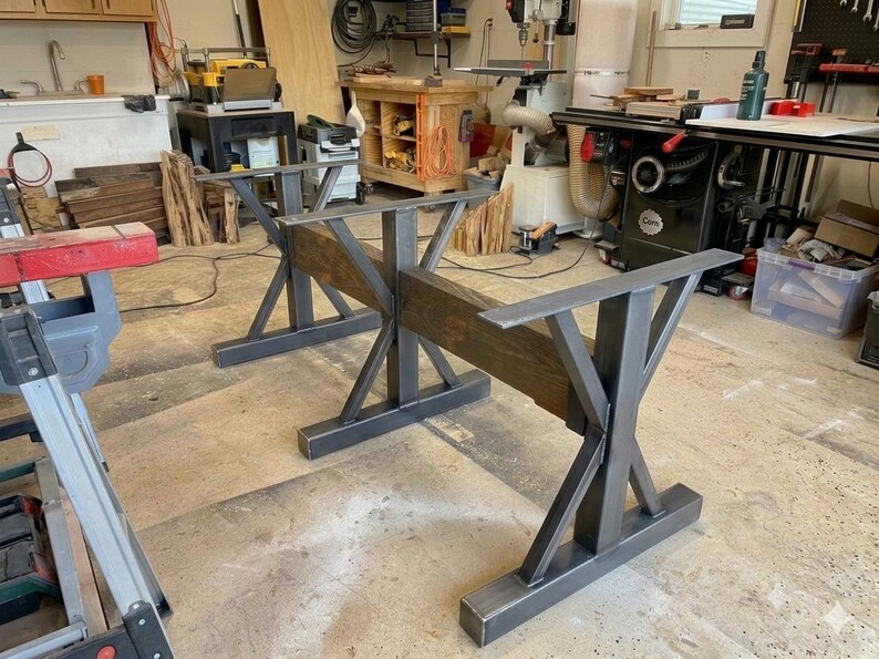 Pu&ograve; includere: Un set di due basi per tavolo realizzate in metallo grigio scuro e legno tinto marrone. Ogni base presenta una struttura di supporto centrale in metallo a forma di X collegata a una trave di legno orizzontale, con piastre metalliche piatte.
